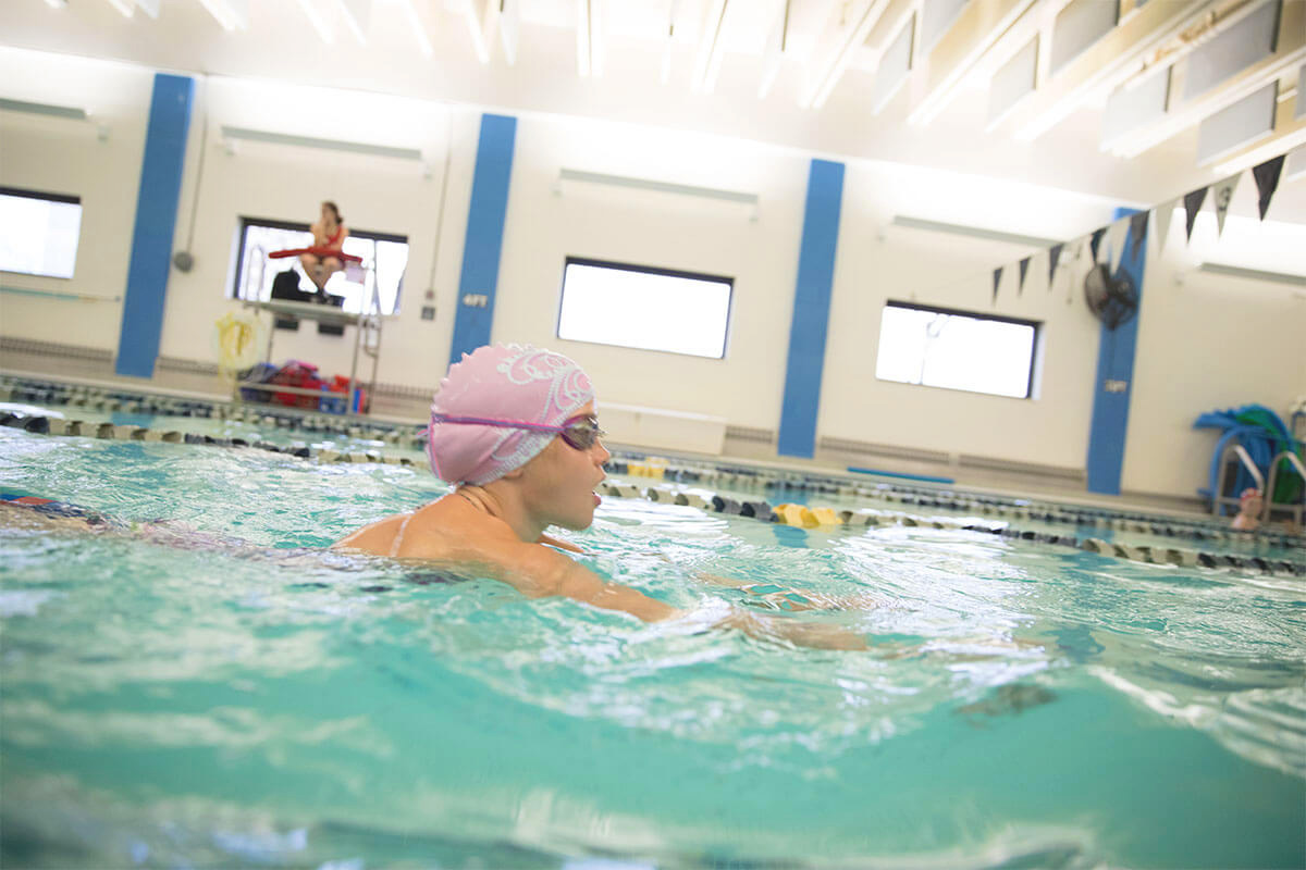 A young girl is exercising in the YMCA swimming pool with goggles and a swim cap.